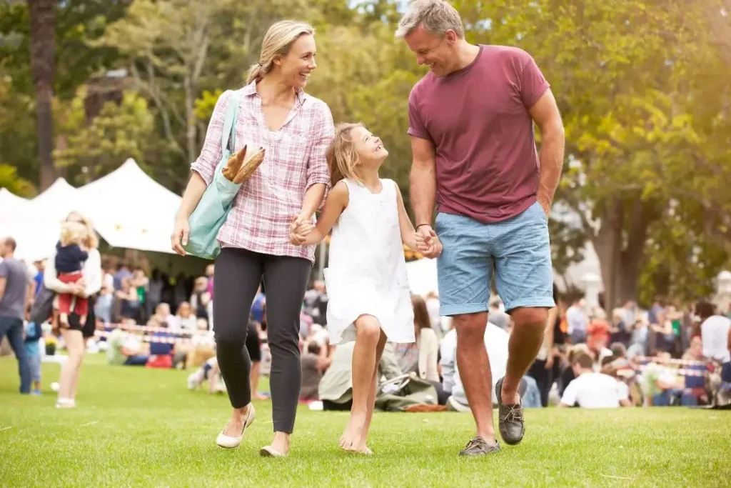 Family of three walking through a crowded sunlit park in Camas, WA, enjoying the Camas Students and Families Foundation fundraiser.
