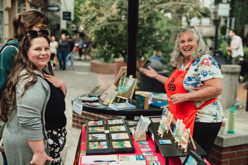 A smiling local artist showcasing framed miniature paintings to visitors at the Camas Art and Wine Walk.