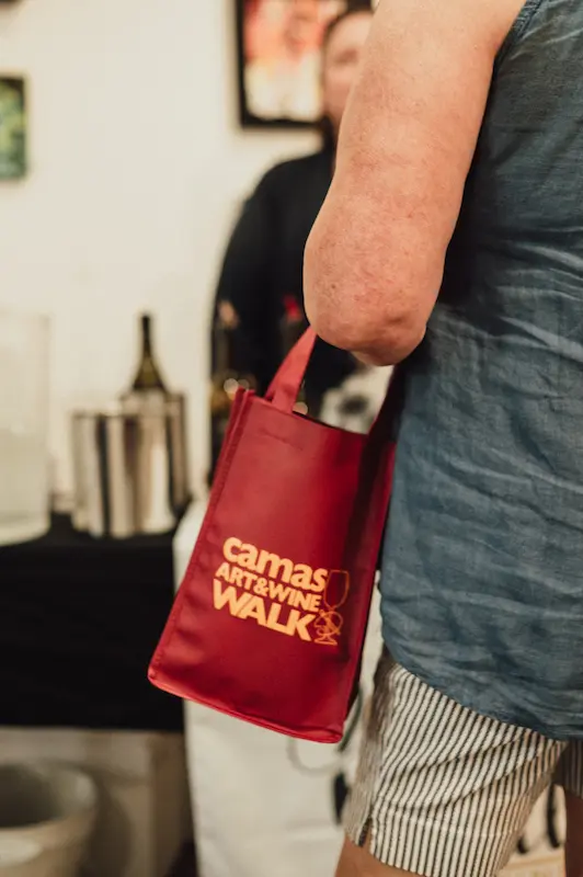 Close-up of a visitor holding an official red Camas Art and Wine Walk reusable bag while exploring the festival.