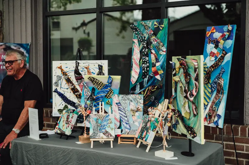 A local artist displaying unique mixed-media necktie paintings at a sidewalk booth during the Camas Art and Wine Walk.